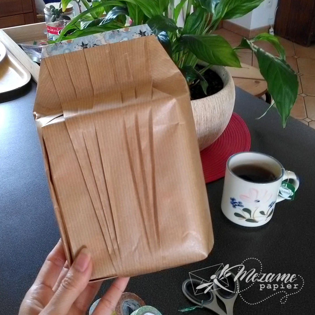 Paquet cadeau en kraft blond d'une boîte de forme non rectangulaire avec des plis selon une technique de l'école Shiho Masuda, avec une décoration de washi tape bleu ciel compportant un motif de grues