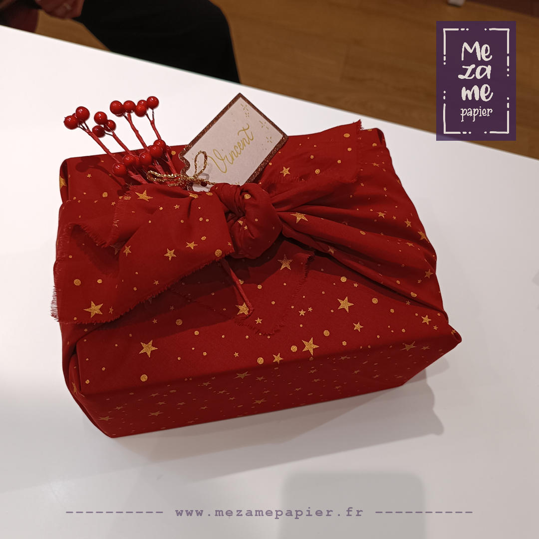 Coffret de cosmétiques de luxe emballé adns du tissu rouge à étoiles jaunes, posé sur une table blanche. Le nœud est horizontal et une branche de baies décoratives factices y est insérée.