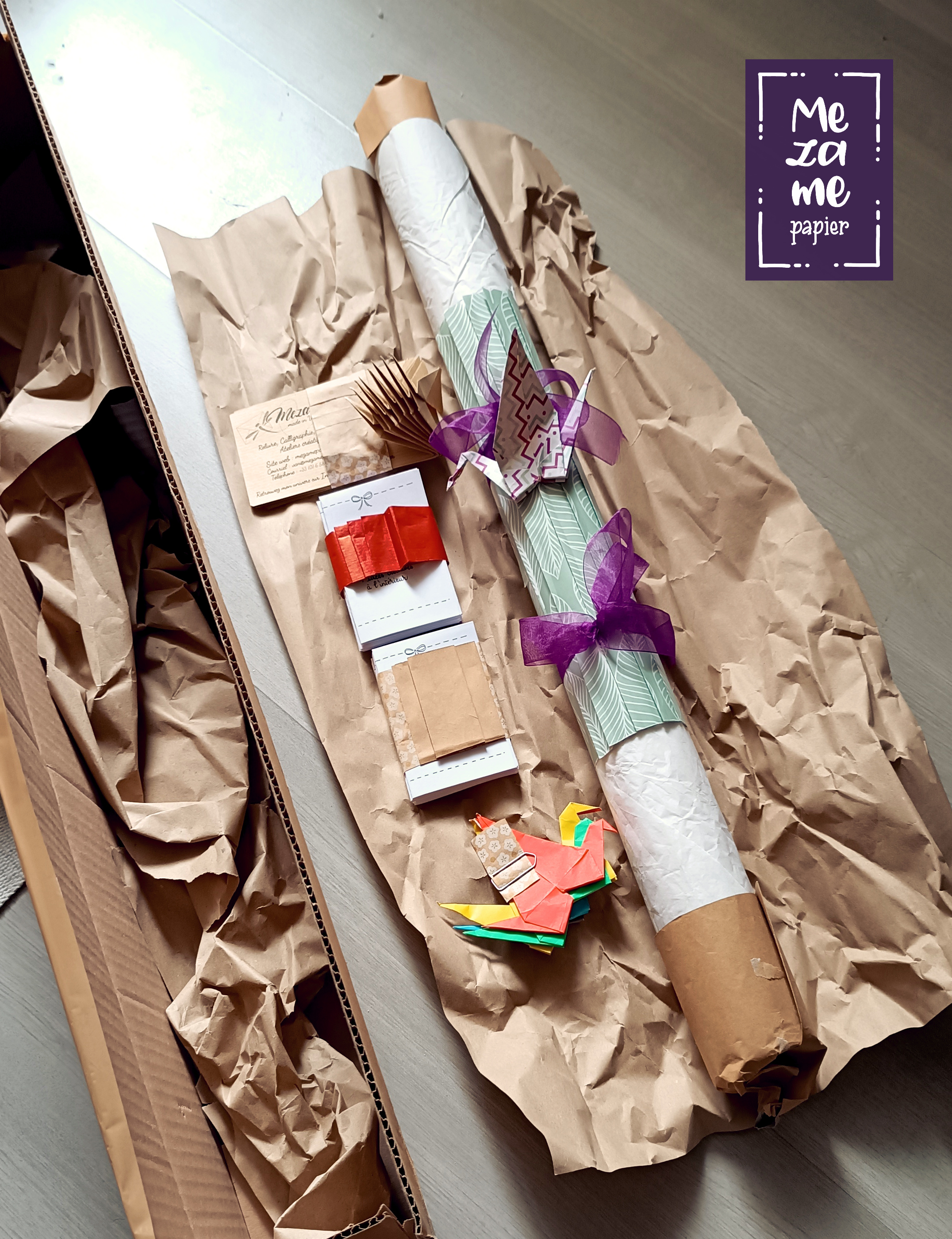Contenu d'un colis envoyé suite à annulation d'un atelier créatif : rouleau de papiers emballé avec soin dans du papier de soie blanc et une ceinture de papier imperméable vert sauge avec des plis décoratifs et 2 rubans d'organza violet vif, cartes de visite en papier kraft brun, mini livrets cadeau retenus par une ceinture de papier rouge, quelques grues en papier origami coloré retenues par un trombone. Les éléments sont posés sur du papier de calage brun et la boîte allongée rectangulaire est ouverte.